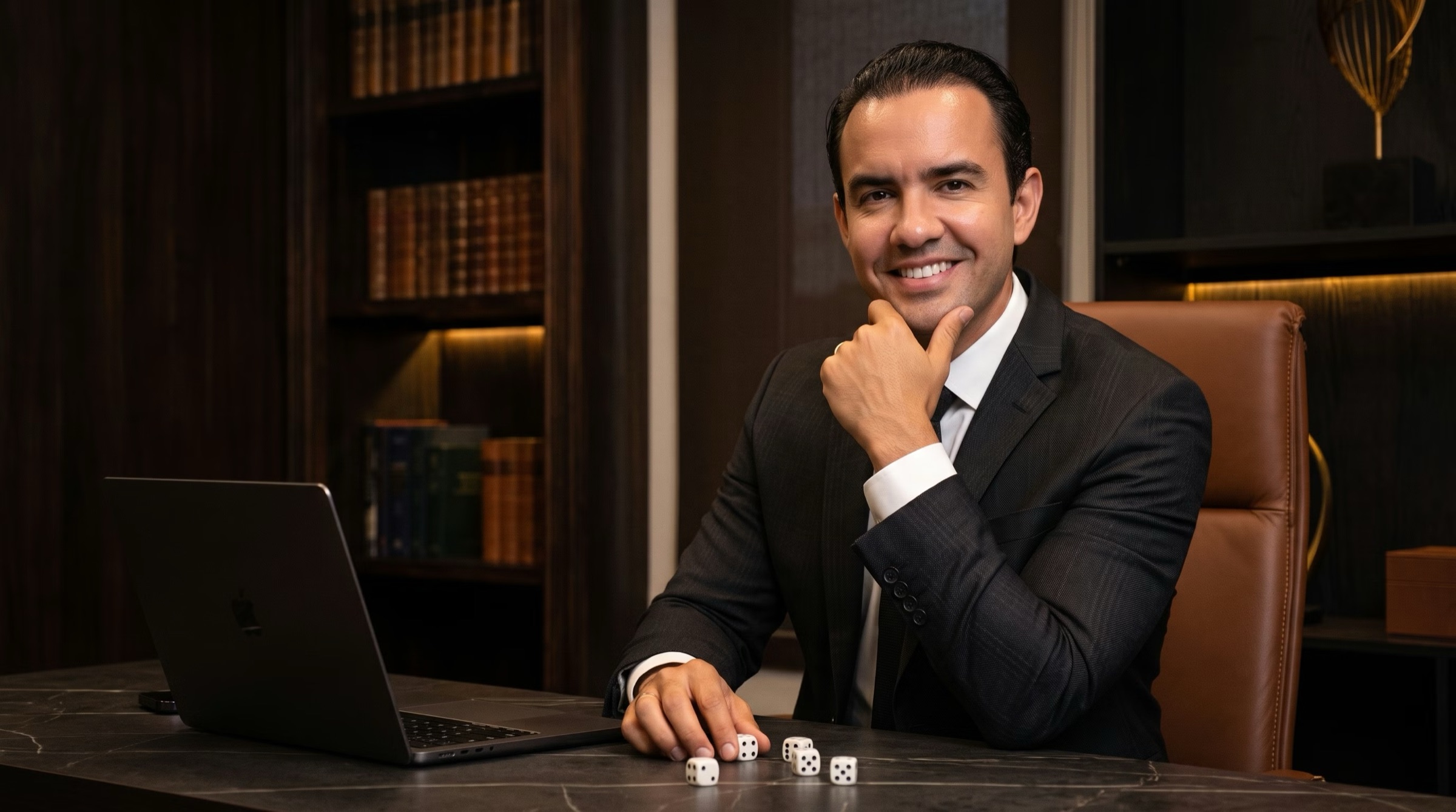 Advogado do escritório Campelo, de terno escuro, sorrindo à mesa de mármore em frente a uma estante de livros jurídicos.